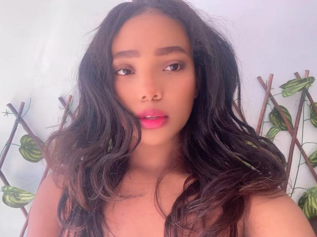 DiannaX69 with long, wavy black hair, brown eyes, pink lipstick, and a cute, sexy face, wearing a sleeveless top, showing her soft, medium-sized tits.