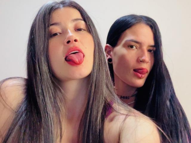 DakottaScarlett in the foreground with long brown hair, red lips, and tongue out, DakottaScarlett in the background with black hair, nose ring, and tongue out.
