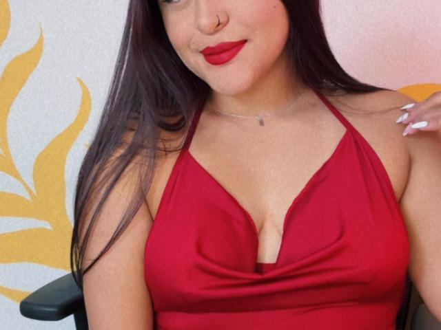 MarianaVargasX with red halter dress, showing her big tits in a deep cleavage, red lipstick, long dark hair, white nails, and a nose ring, sitting on a chair.