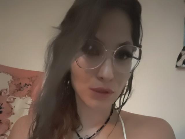 Adilay, avec ses cheveux bruns ondulés, portant des lunettes rondes et un haut blanc à encolure dégagée, montrant son visage et sa petite poitrine, légèrement floue, avec un sourire discret..