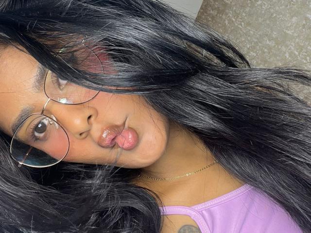 Babilett with long black hair, puffy pink lips, and glasses, wearing a purple top, has a small tattoo on her shoulder, and a delicate necklace.
