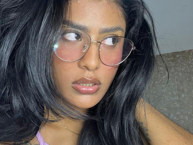 Babilett has dark, wavy hair, pink-tinted glasses, and glossy pink lips. Her brown skin is slightly shiny, and she's wearing a thin gold necklace.