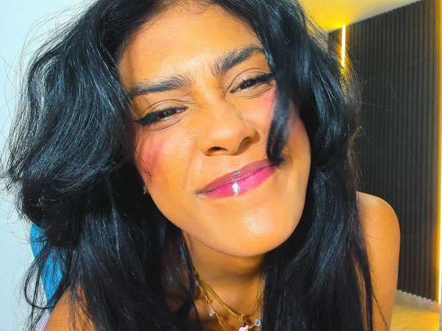 Babilett with dark, wavy hair, pink lipstick, and a slight smile, showing off her glossy pink pout and relaxed eyes. She's wearing a gold necklace.