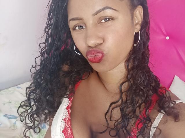 LayahCoquine with curly black hair, red lipstick, hoop earrings, and a red lace bra, pouting her lips, showing her boobs, and looking at the camera.