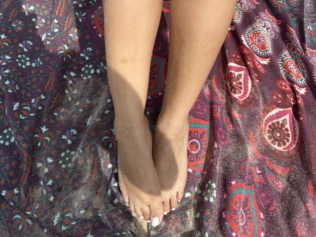 NashiroSakuraii's feet are crossed at the ankles, with sandy skin and white-painted toenails, standing on a colorful, patterned cloth. Her toes are slightly curled.