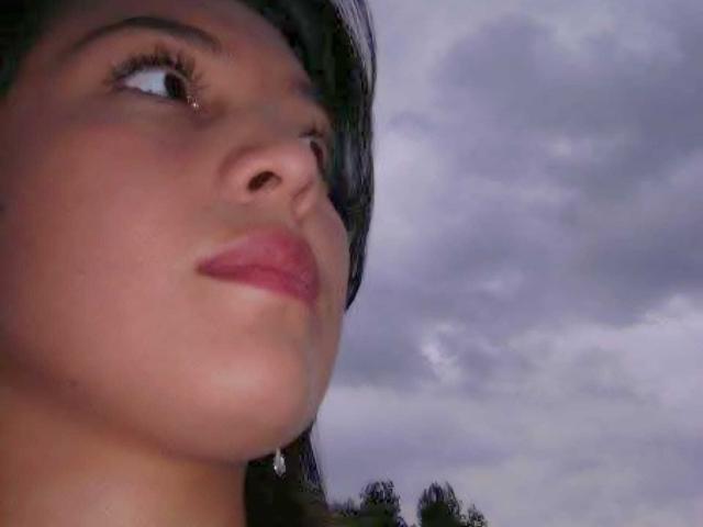 PaulaAnd's face, with smooth tan skin, pink lips, and dark eyelashes, looking up at the cloudy sky, wearing a single dangling earring on the left side.