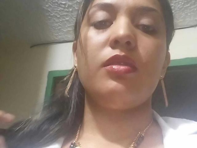 PaulaAnd has shiny brown skin, glossy red lips, and dark straight hair in a ponytail, wearing gold earrings and a necklace, looking up with a slightly pouty expression.