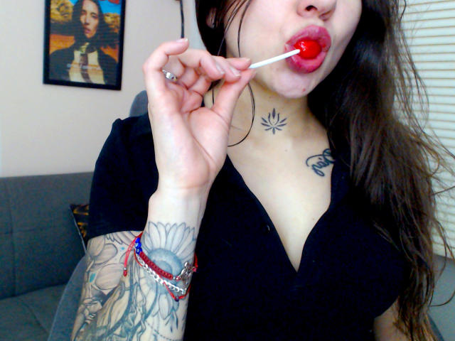 SujannaVarma with long brown hair, black shirt, red lollipop in mouth, tattoos on neck and arm, sunflower tattoo, holding lollipop near lips, showing off tattoos and jewelry.