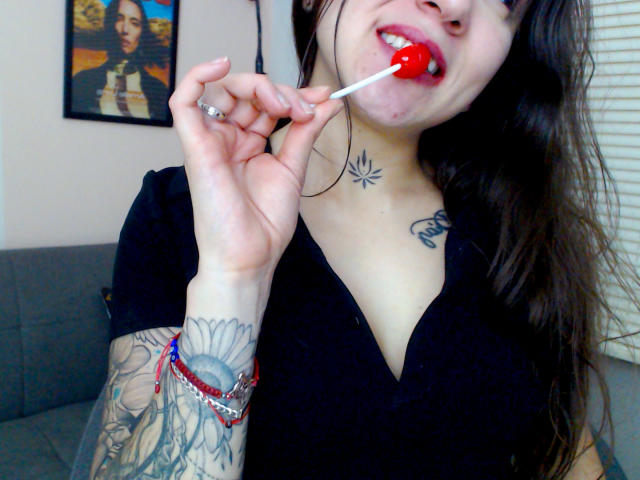 SujannaVarma with tattoos on her arm and chest, wearing a black V-neck, sucking on a red lollipop, showing her puffy pink lips. She has long brown hair.