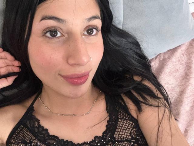 SophieBeaumont has black lace lingerie on, with long dark hair, brown eyes, and pink lips, lying on a bed, wearing a thin chain necklace.