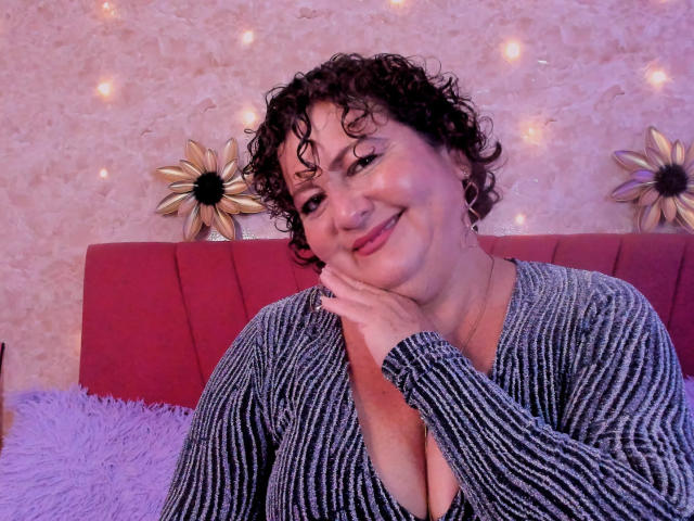 Curly-haired PalomaLombardi with a cute smile, resting her chin on her hand, wearing a striped grey shirt, showing her boobs, purple pillow, and red headboard.