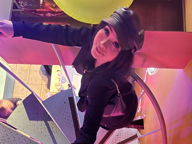 NashiroSakuraii, wearing a black leather cap and jacket, leans on a metal railing with a playful smile, showing her pouty lips and dark makeup, on a staircase.