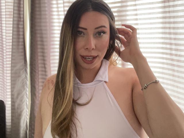 JennyLawry with long brown hair, white halter top, brown eyes, and a bracelet, smiling and touching her hair, showing her tits.