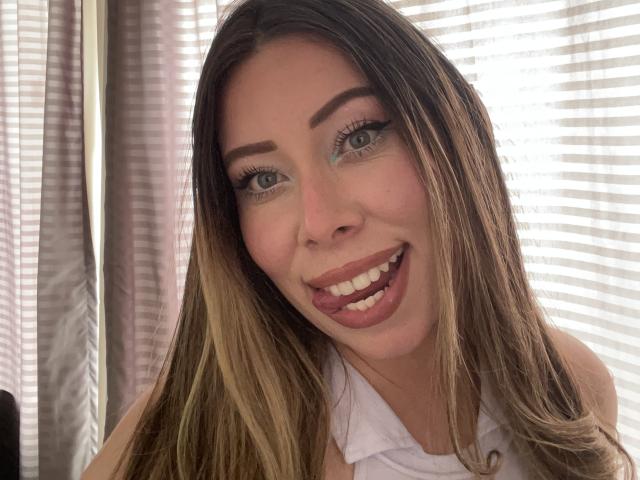 JennyLawry with long brown hair, green eyes, and a big smile, sticking out her tongue, wearing a white shirt, showing her teeth and a cute face.