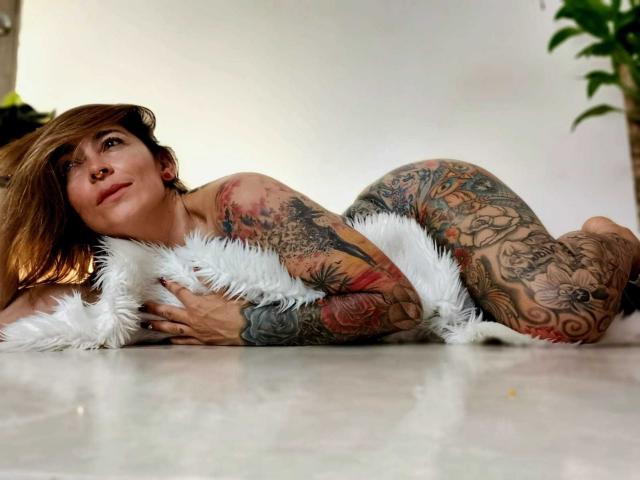 SaraValkyria, nuda, sdraiata su una coperta bianca e soffice, con tatuaggi che coprono braccia e glutei, capelli castani, piercing al naso, che guarda di lato, in una stanza con una pianta verde..