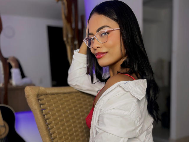ChloeBrownOne with long black hair, red lingerie, white shirt, and glasses, smiling at camera, hand on chair, wicker chair, bedroom, blue light, sexy, attractive, confident.