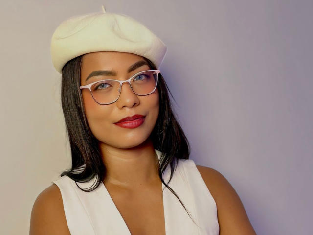 ChloeBrownOne has dark brown skin, wearing a white beret, pink glasses, and red lipstick, with straight black hair, and a white sleeveless top, smiling gently.