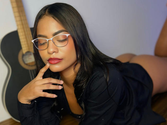 ChloeBrownOne with long black hair, wearing glasses and a black shirt, lying on her stomach, showing her ass, with a finger near her red lipstick, and a guitar.