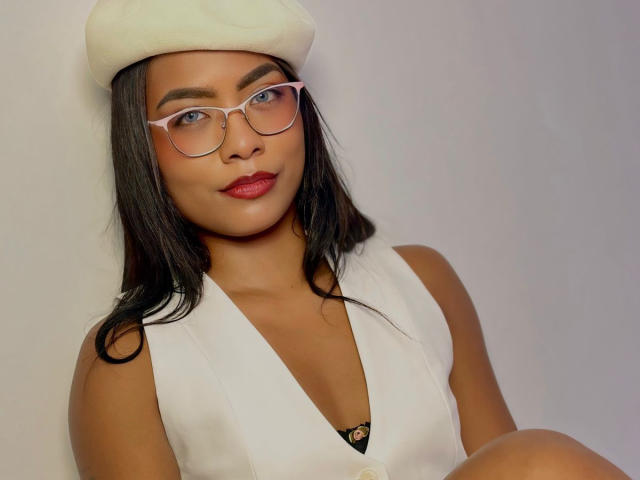 ChloeBrownOne with dark hair, red lipstick, white beret, and glasses, wearing a white vest, showing a black bra, her tits slightly visible, sitting with a relaxed smile.