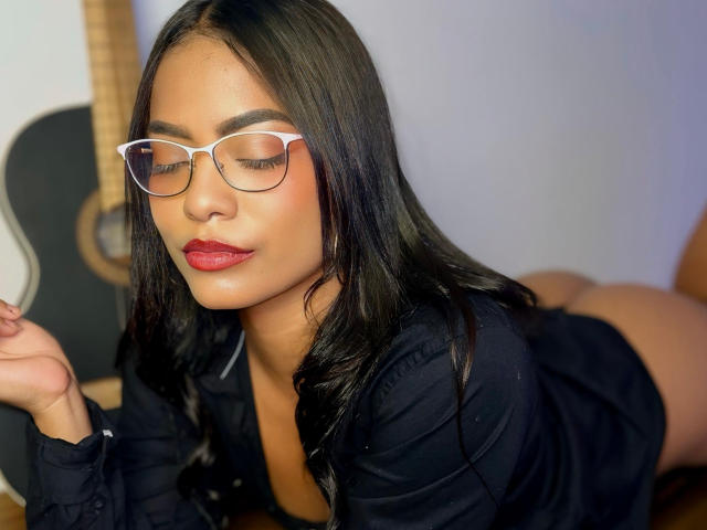 ChloeBrownOne with black hair, red lipstick, and white glasses, lying on her stomach, wearing a black shirt, showing her ass, looking down with a relaxed expression.