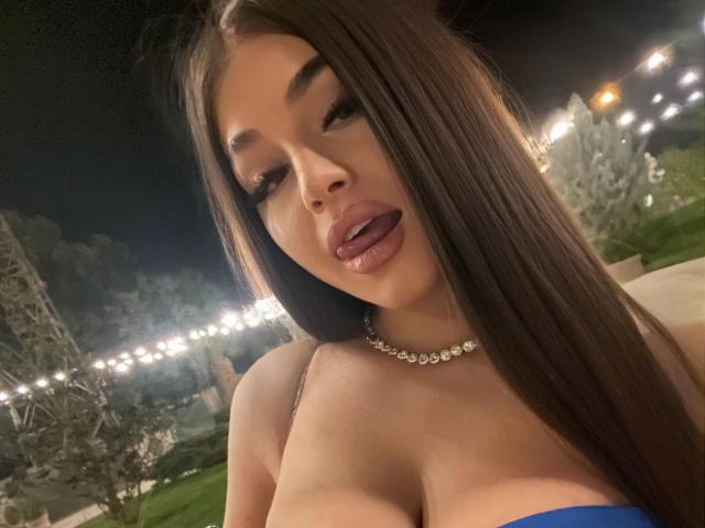 BellaNoirre ha lunghi capelli castani, labbra piene e sta mostrando la lingua. Indossa un top blu senza spalline che mette in mostra il suo seno prosperoso, e una collana di diamanti..