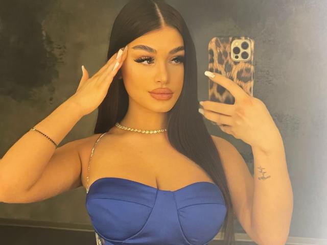 BellaNoirre with long black hair, blue strapless dress, big tits, gold necklace, leopard phone case, holding mirror, saluting, white nails, tattoo on right wrist, golden skin.