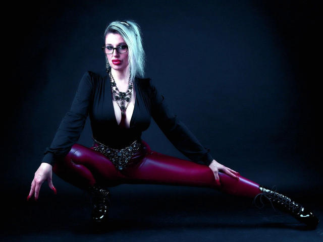 Avengelique-hot with pale skin, red lipstick, and glasses, squatting in a black top and red leather pants, showing her tits and ass. Wearing a black necklace and black boots.