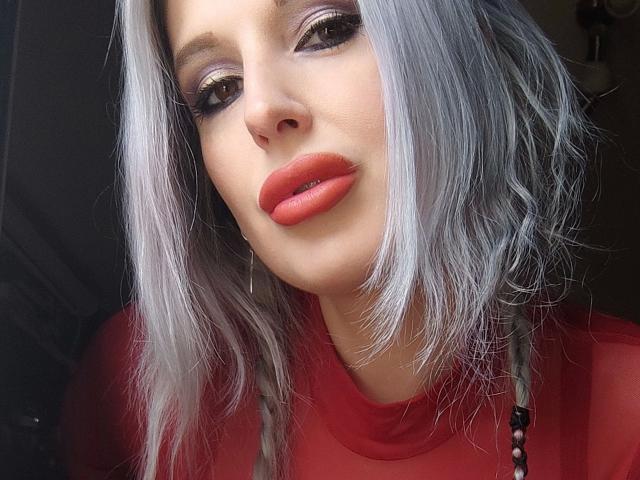 Avengelique-hot has silver hair, red lipstick, and wears a red shirt. She has purple eye makeup, and her tits are slightly visible under the shirt.