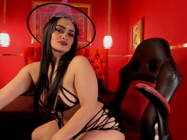 AnissaAli with long black hair, dark lipstick, wearing a black strappy lingerie set and a hat, sitting on a black chair, red room, sexy, large tits, pale skin.