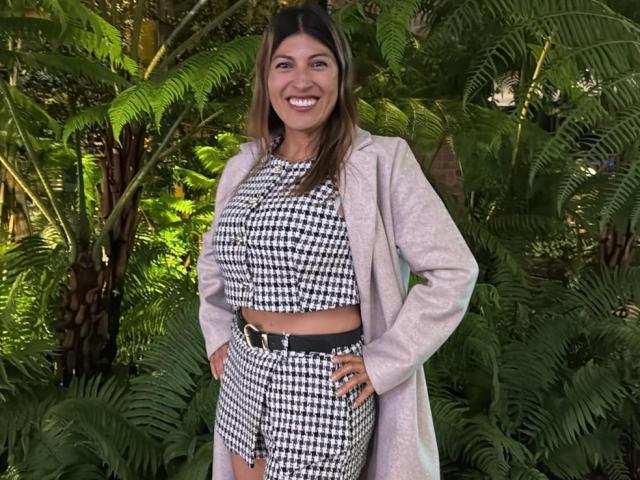 LunaBeya stands smiling, wearing a houndstooth crop top and matching shorts, with a pink coat, hands on hips, green ferns in the background, showing her tits and ass.