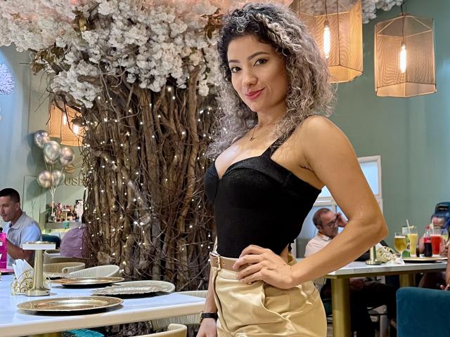 NicolBlosson has curly gray hair, wearing a black tank top showing small tits and gold high-waisted pants, standing in a cafe with a floral wall and hanging lights.
