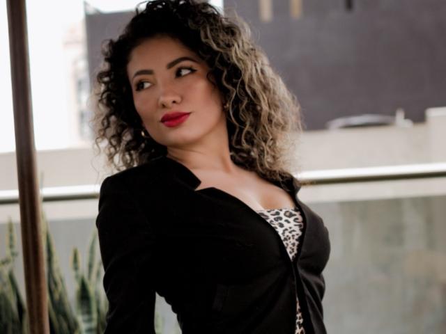 NicolBlosson with curly gray and black hair, red lipstick, black cardigan, and leopard print top, looking to the side with a slight smile. Boobs visible under cardigan.