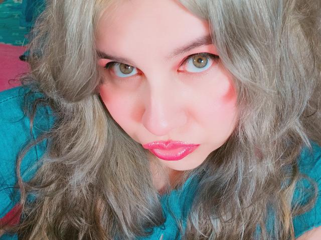 Het gezicht van Akkanee, met golvend grijs haar, roze lippen en rode oogmake-up, kijkt recht in de camera met groene ogen. Ze draagt een blauw shirt..