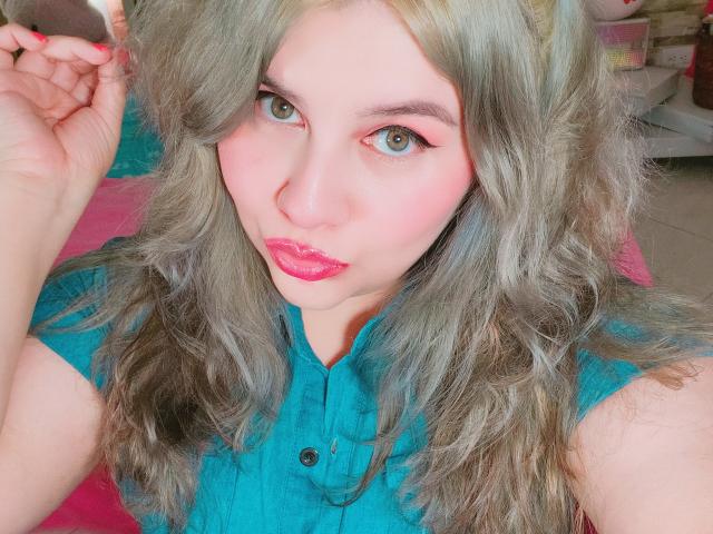 Akkanee with pale skin, green eyes, pink eyeliner, and glossy pink lips, wearing a teal shirt, has wavy grayish-blonde hair, and a slightly pouty expression, raising her hand.