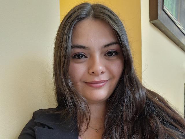 KatherinLatin has long brown hair, brown eyes, and is smiling with her teeth slightly showing. She's wearing a black jacket over a pink shirt, and a gold necklace.