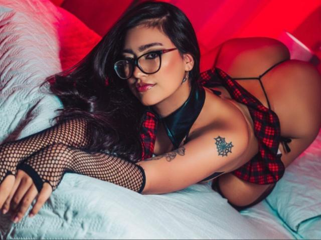 TatuadaSafadaHot with long blonde hair, tattoos on arms and side, topless, black jacket, black thong, sitting on chair, looking back, showing tits, tattoos, and a sexy face.