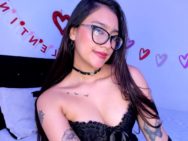 AgathaHowlandd with long black hair, glasses, black lace corset, sheer gloves, and black thigh-highs, showing her butt, tattoo on right shoulder, kneeling on bed with pink flowers.