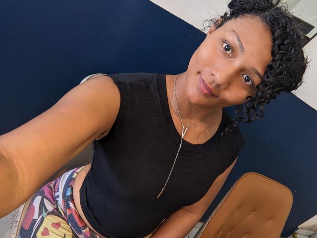Ninah69s with curly black hair, wearing a black sleeveless top, necklace, and colorful patterned pants, smiling at the camera, holding her phone for a selfie.