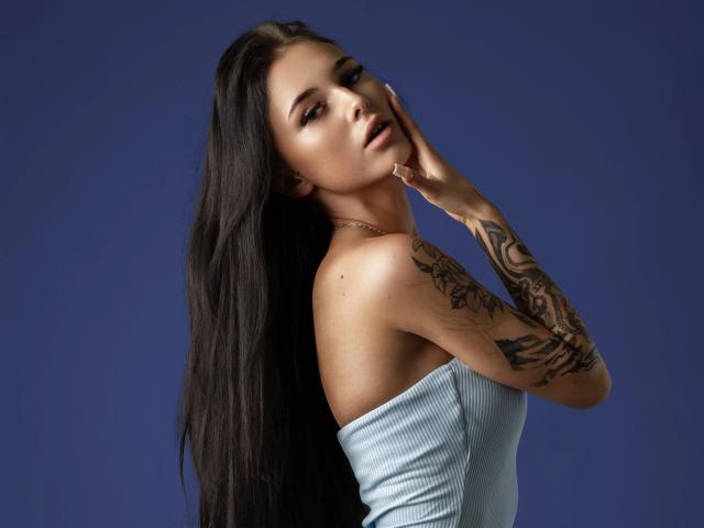 Oliviaelika-hot with long black hair, tattooed arm, wearing a strapless light blue top, hand on cheek, blue background, looking seductively at the camera.