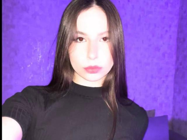 Amabel has long black hair, pale skin, and red lips. She's wearing a black top, looking at the camera with a neutral expression, in a purple-lit setting.