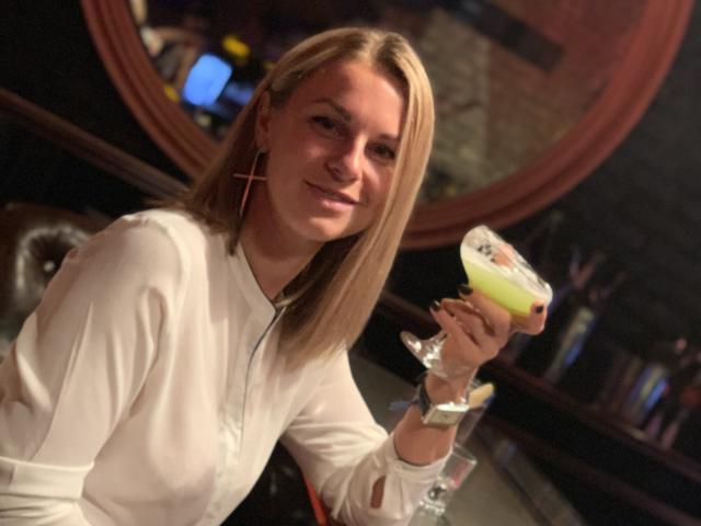 BelleLisaG with blonde hair, white shirt, and cross earrings, holding a green cocktail in a glass, smiling slightly, sitting at a bar.