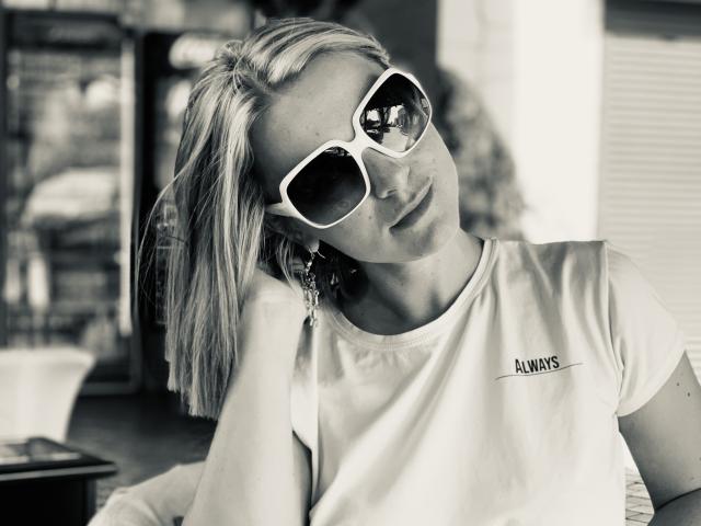 BelleLisaG mit blonden Haaren, weißen Sonnenbrillen mit weißem Rahmen und einem weißen "Alumis"-T-Shirt, die auf ihrer Hand gestützt ist und leicht zur Seite schaut. Schwarz-weiß-Foto, leichtes Lächeln..