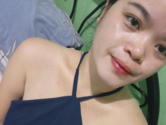 OpenMindedKinyStar with fair skin, black hair, and small tits, wearing a navy halter top, smiling softly at the camera, lying on a green bed with a blue pillow.