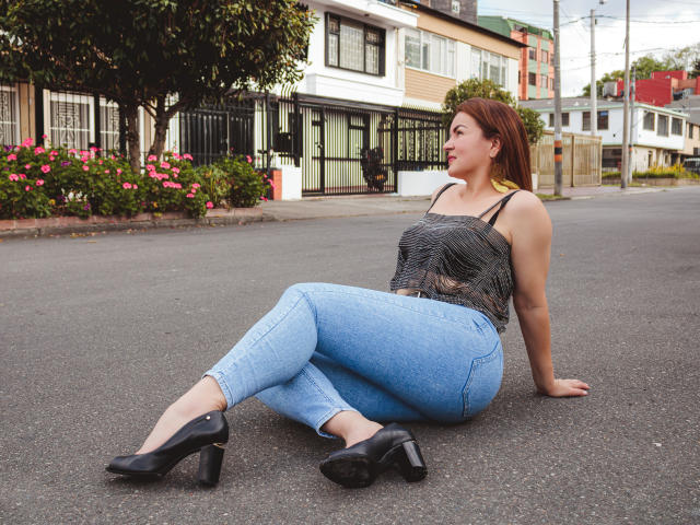 VictoriiaJones com cabelo ruivo, blusa preta estampada, jeans azuis, saltos altos pretos, sentada no asfalto, pernas cruzadas, braços atrás, olhando para o lado, nádegas levantadas, seios visíveis..