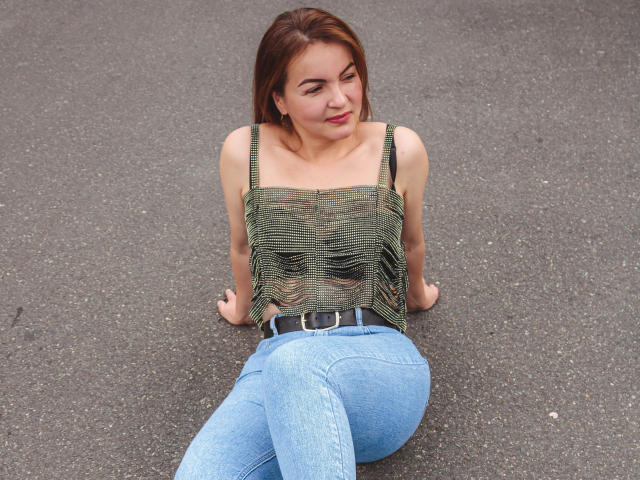 VictoriiaJones, com cabelo castanho, blusa verde listrada e calça jeans azul, sentada em asfalto cinza, com os braços atrás, olhando para baixo, com o bumbum ligeiramente levantado, cinto visível, sorrindo suavemente..