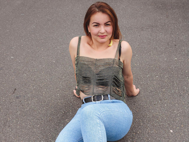 VictoriiaJones met een gemiddelde borstmaat, zittend op asfalt, gekleed in een gestreepte tanktop, blauwe jeans en een zwarte riem, lachend, met bruin haar en gele oorbellen..