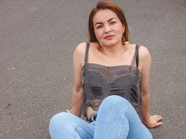 VictoriiaJones está sentada no asfalto, vestindo uma camiseta listrada preta e branca, jeans azuis e brincos amarelos, com um leve sorriso, e seu cabelo castanho solto..