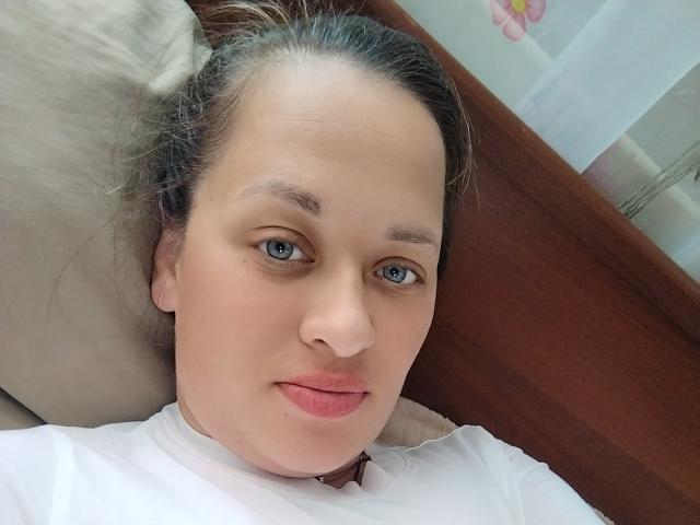 MarleneFan with blue eyes, light skin, and pink lips, lying on a pillow, wearing a white shirt, has brown hair pulled back, and a slightly relaxed expression.
