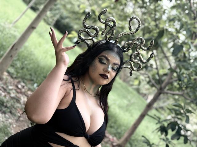 Lara James con un tocado con forma de serpiente, maquillaje negro, sombra de ojos verde y lápiz labial oscuro, vistiendo un sujetador negro, mostrando sus grandes senos, levantando una mano, en un bosque..