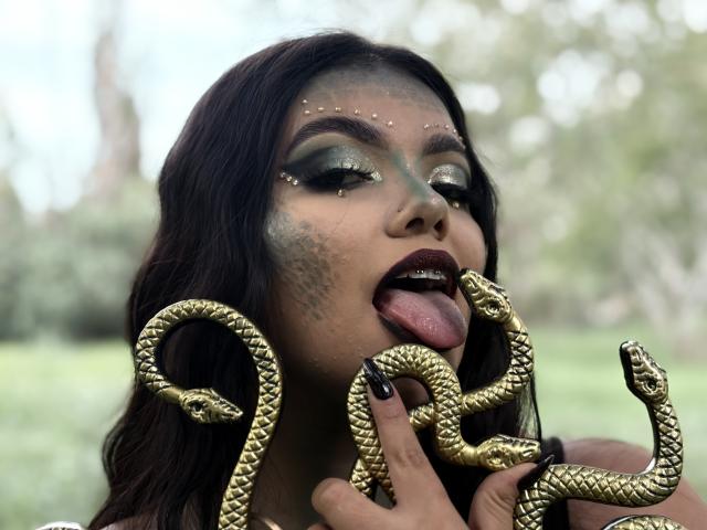 LaraJames with dark hair, green eyeshadow, and gold snake earrings, tongue out, touching gold snake hands, black nails, in a green outdoor setting.